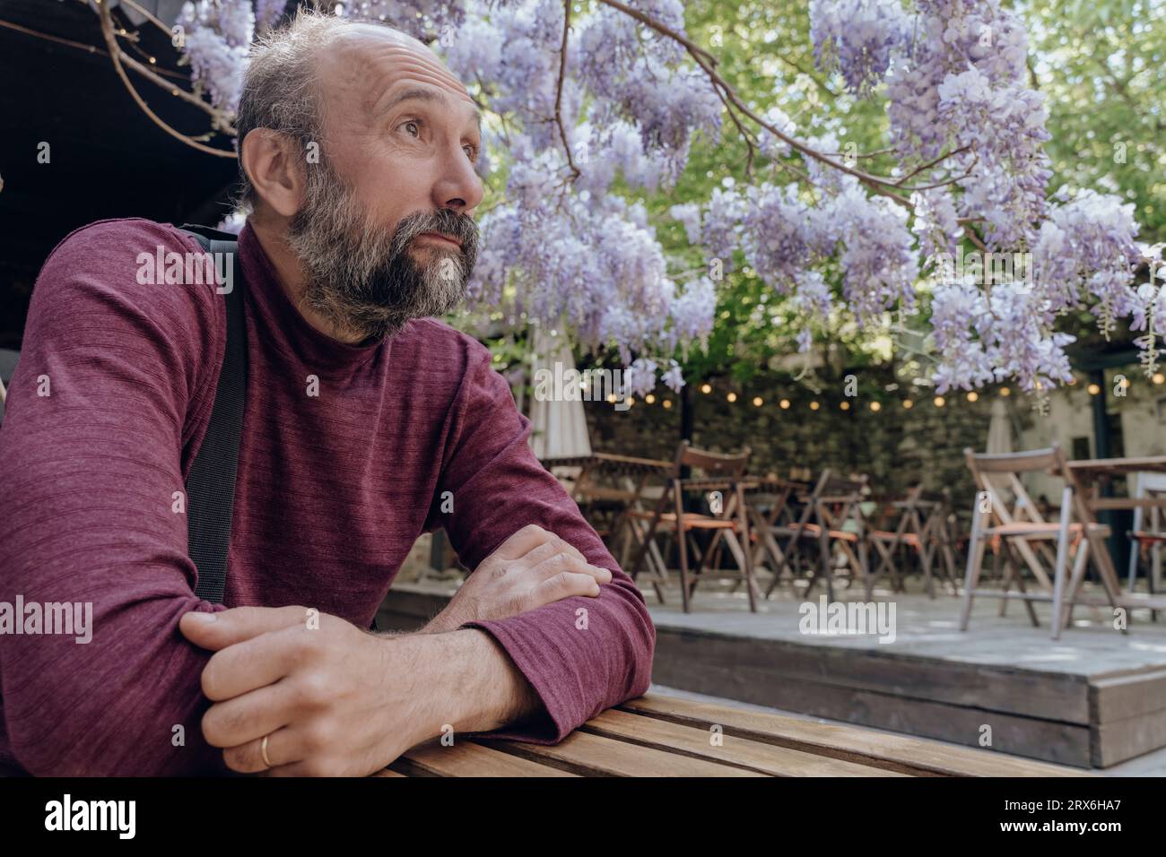 Uomo premuroso seduto con le braccia incrociate sotto l'albero di glicine al caffè sul marciapiede Foto Stock