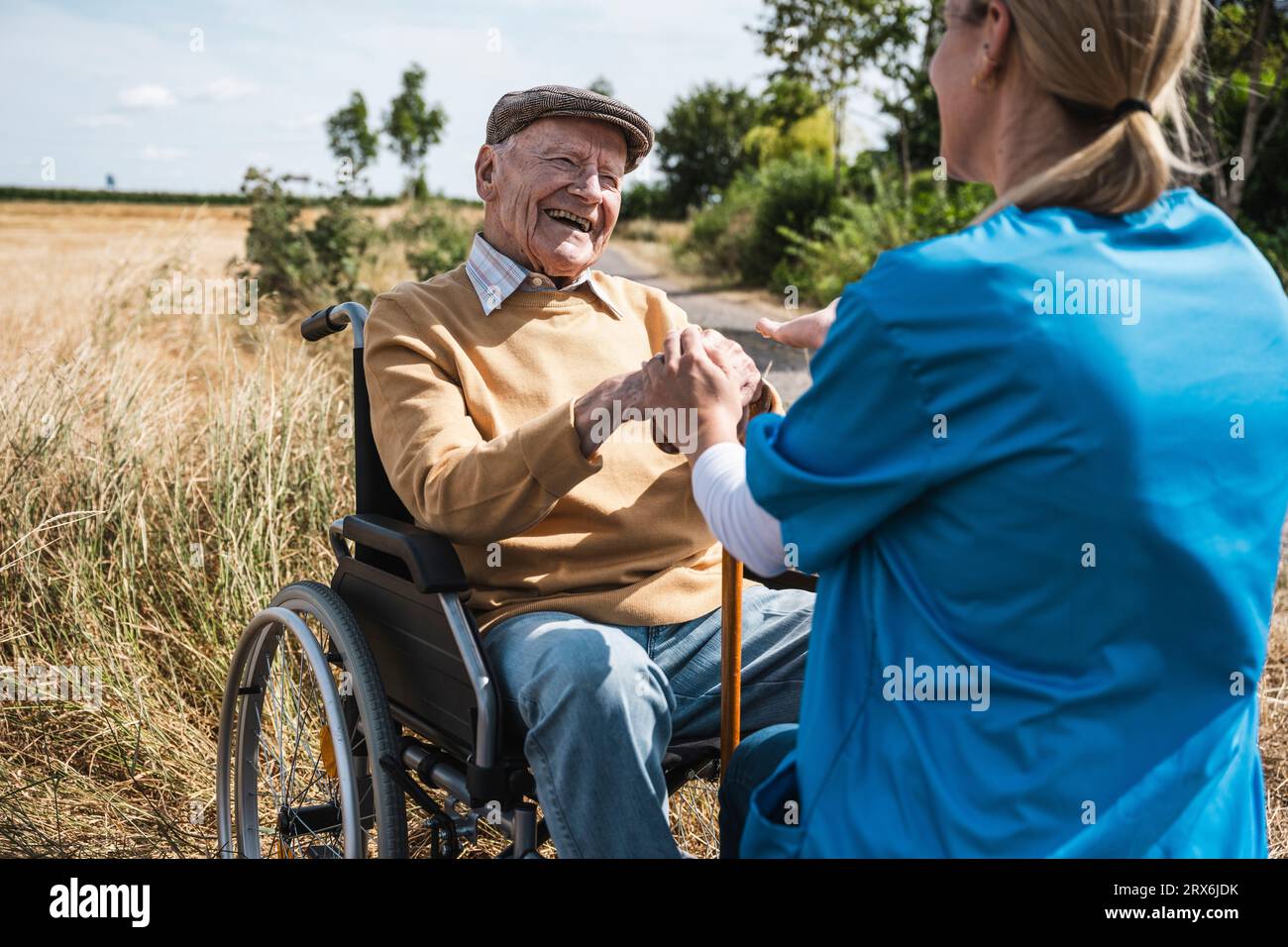 Uomo anziano felice seduto in sedia a rotelle e che parla con l'infermiera durante la giornata di sole Foto Stock