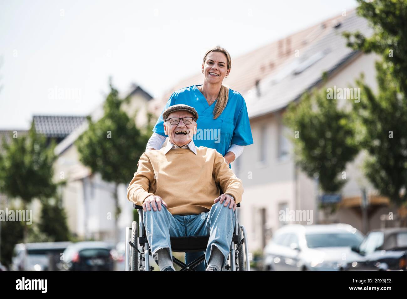 Un uomo anziano allegro seduto in sedia a rotelle e un'infermiera che lo spinge nelle giornate di sole Foto Stock