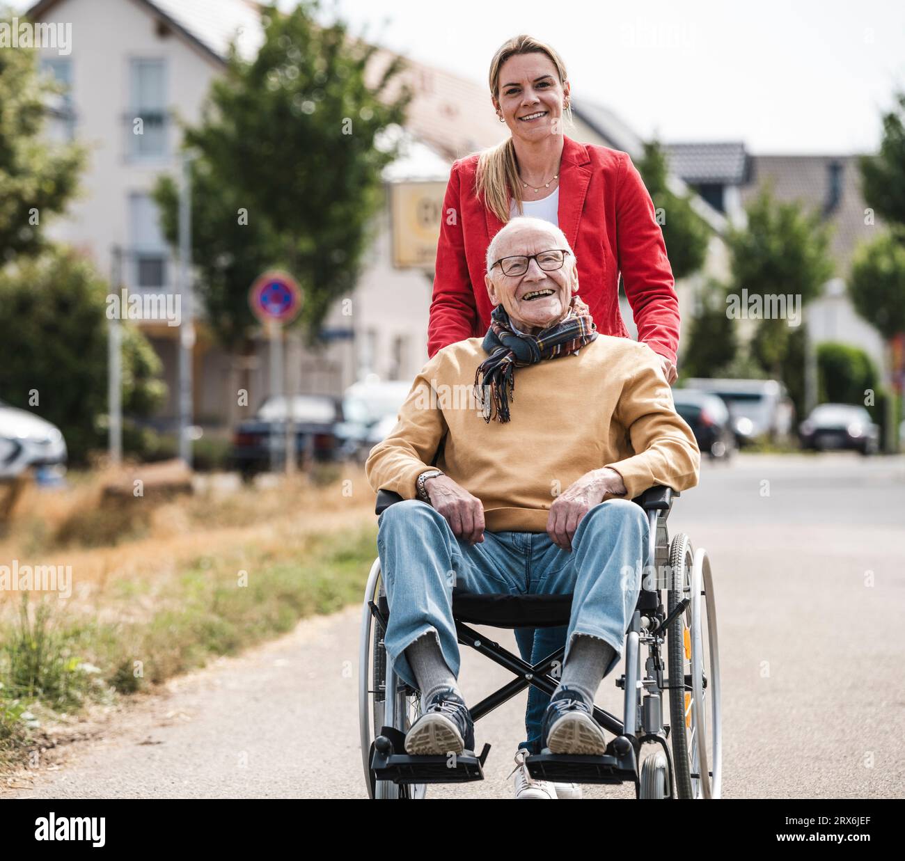 Donna sorridente che spinge un uomo anziano seduto in sedia a rotelle su strada Foto Stock