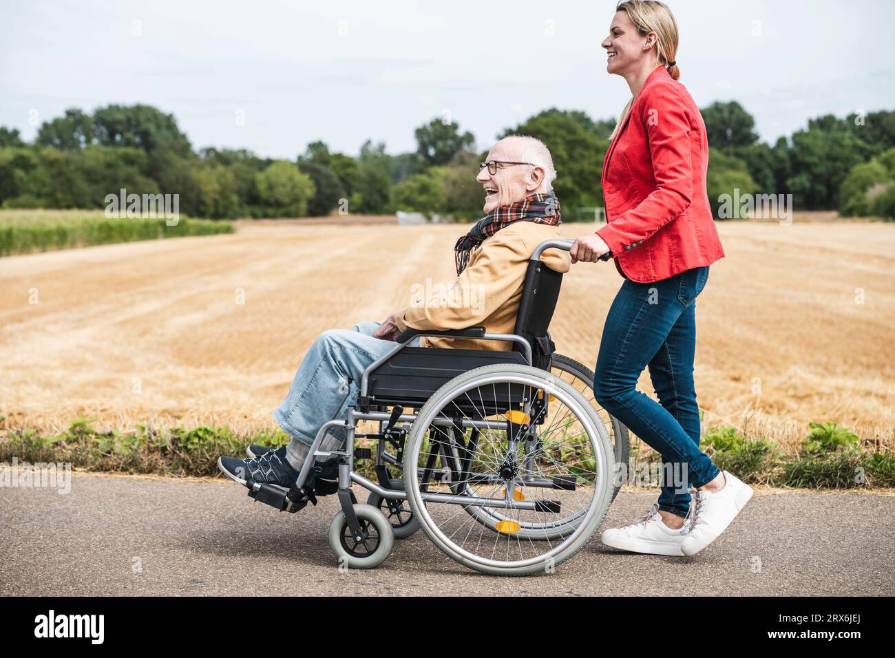 Donna che spinge un uomo anziano felice seduto in sedia a rotelle su strada Foto Stock