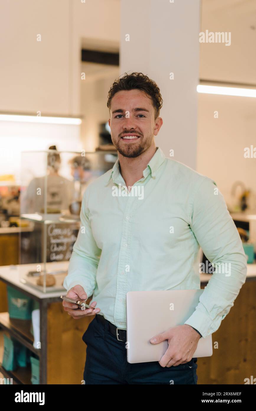 Uomo d'affari sorridente con smartphone e computer portatile in caffetteria Foto Stock
