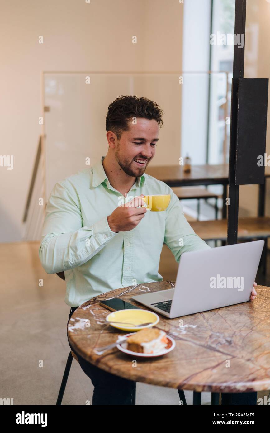 Un uomo d'affari felice con una tazza di caffè che lavora al computer portatile al tavolo del bar Foto Stock