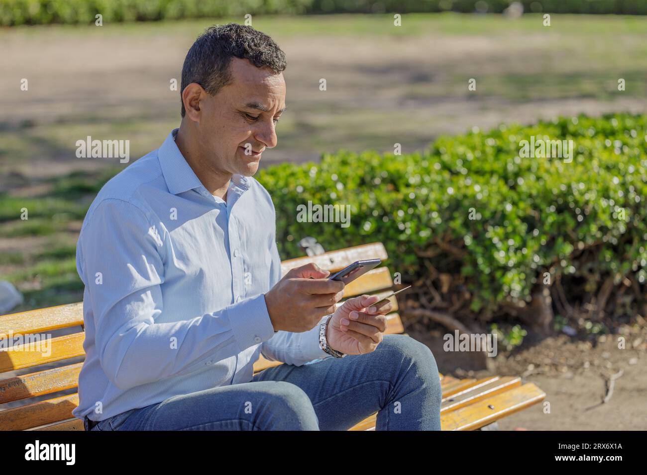 Un giovane latinoamericano che fa un acquisto con una carta di credito sul suo cellulare in un parco pubblico. Foto Stock