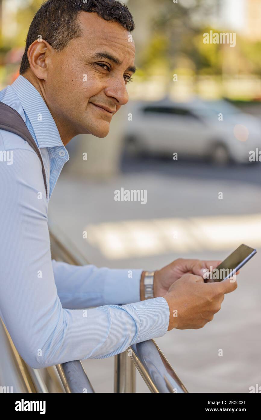 Ritratto di un uomo latino in camicia appoggiato su una ringhiera usando il suo cellulare. Foto Stock