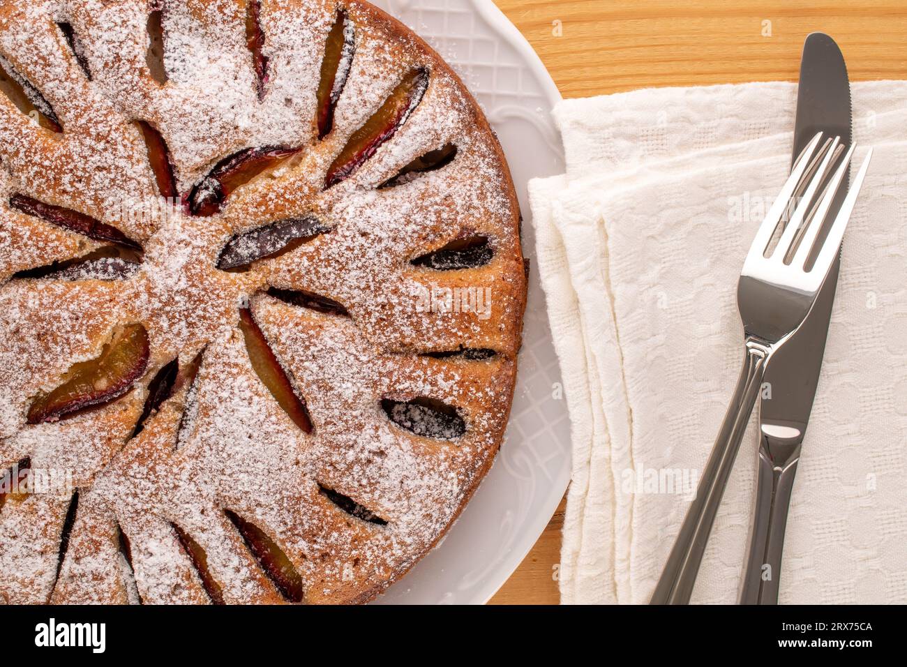 Dolce torta di prugne fatta in casa con piatto in ceramica bianca, coltello e forchetta su tavolo in legno, macro, vista dall'alto. Foto Stock