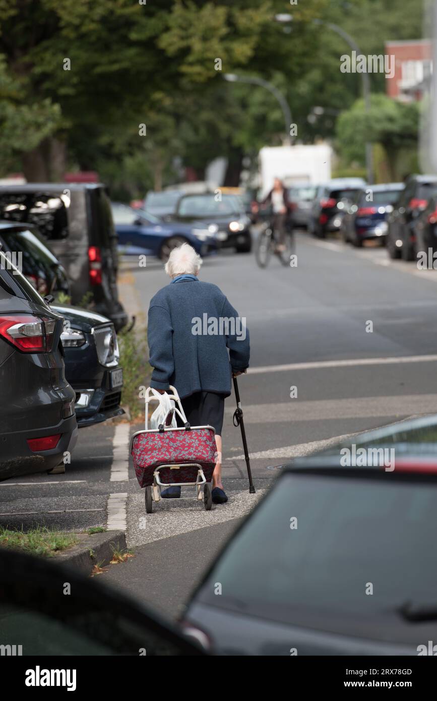 Vecchia donna con camminatore sulla strada accanto al SUV e ad altre auto nel parcheggio degli ospiti. Il ciclista si allontana dall'auto per evitare incidenti. Foto Stock