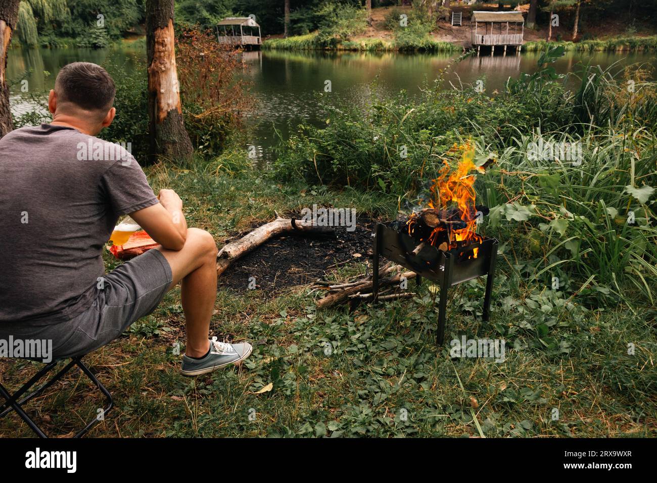 Uomo seduto nella foresta vicino al falò. Uomo in un picnic autunnale. Uomo solitario sulla riva del fiume con fuoco da campeggio. Turismo nei boschi. Turista vicino al caminetto sulla riva dello stagno Foto Stock