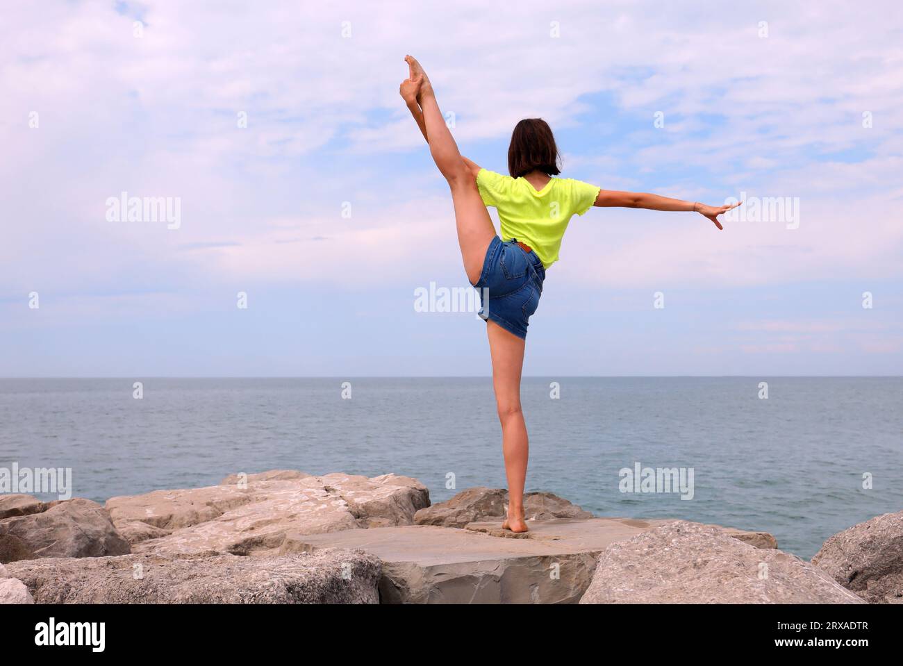 giovane ragazza snella che lancia la gamba in aria durante gli esercizi di ginnastica sulle rocce in riva al mare Foto Stock