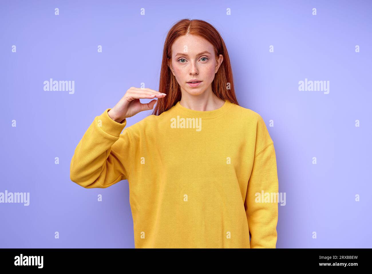 Bella donna rossa seria sicura di sé in maglione giallo che mostra il simbolo della lettera D del linguaggio dei segni per gli umani sordi con sfondo blu. isolato Foto Stock