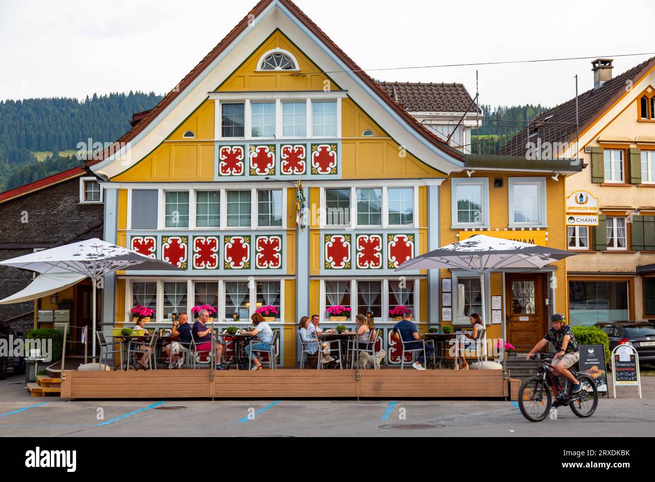 Un ciclista cavalca davanti ai commensali all'aperto all'esterno di questo edificio in stile Appenzeller che ospita il ristorante Vietnamita Chanh ad Appenzell, Svizzera. Foto Stock