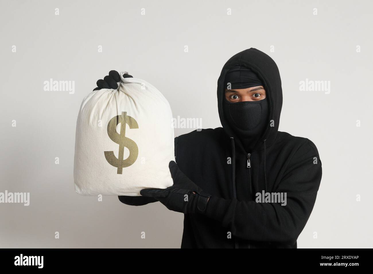 Misterioso ladro di rapinatori che indossa una felpa nera con cappuccio e una maschera e porta con sé una borsa piena di soldi. Immagine isolata su sfondo grigio Foto Stock