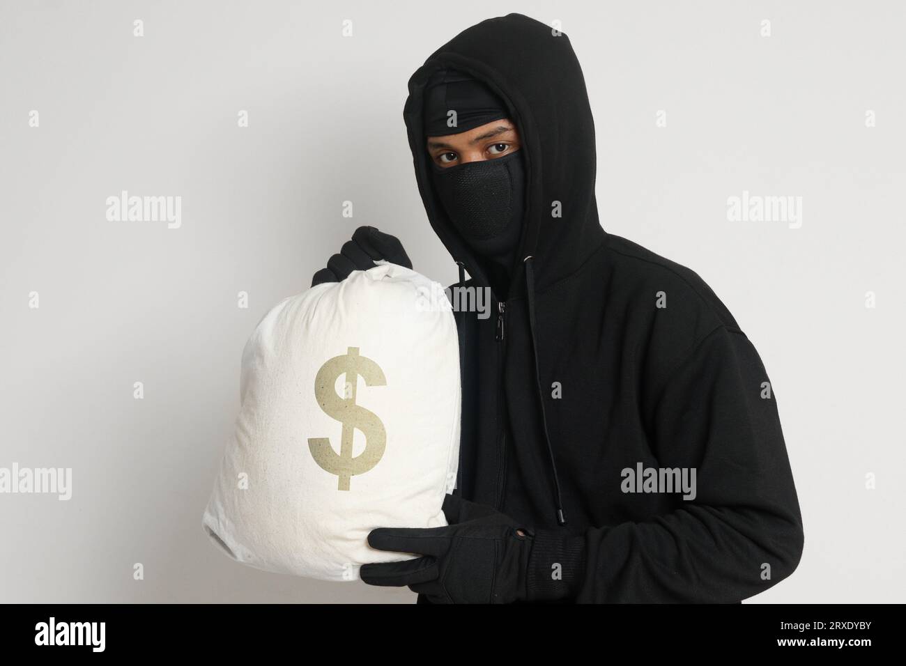 Misterioso ladro di rapinatori che indossa una felpa nera con cappuccio e una maschera e porta con sé una borsa piena di soldi. Immagine isolata su sfondo grigio Foto Stock