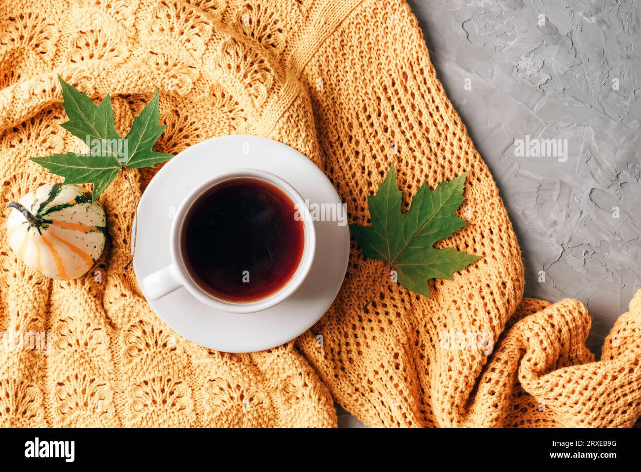 Piano autunnale con foglie verdi, zucca e tazza di caffè su un maglione giallo su sfondo grigio in cemento. Vista dall'alto, spazio di copia. Foto Stock
