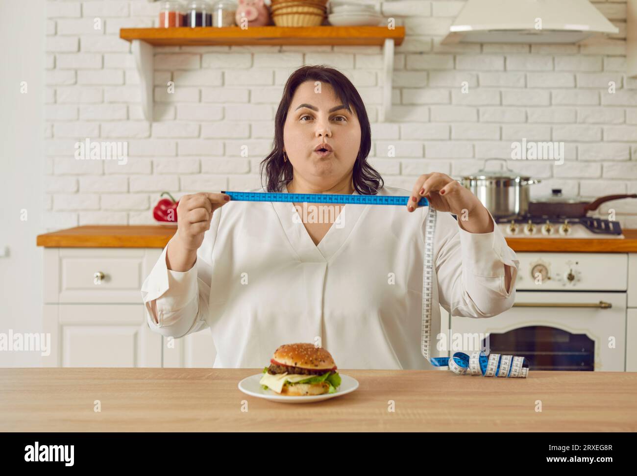 Donna grassa e sovrappeso seduta al tavolo con hamburger e con un metro a nastro Foto Stock