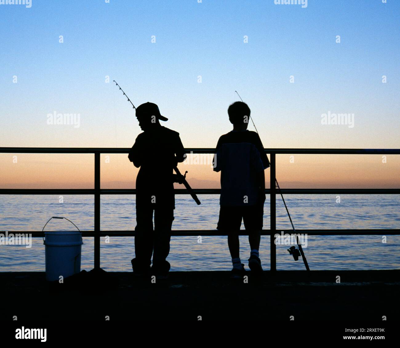 Vista posteriore di due ragazzi con bastoncini da pesca sagomati su un molo al crepuscolo. California. Foto Stock