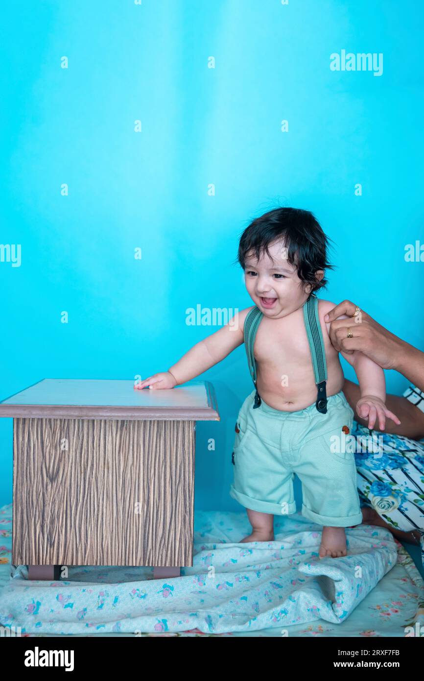 Sorridente bambino indiano di sei mesi che cerca di stare in piedi con il sostegno di sgabello di legno mentre la madre tiene isolata sullo sfondo blu dello studio, ha Foto Stock