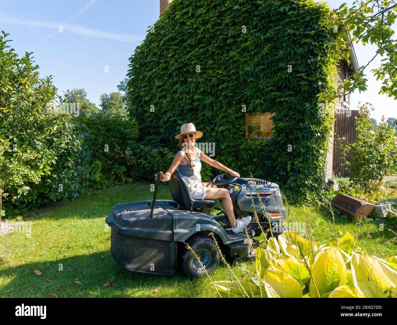 Donna che guida rasaerba, trattore, cappello di paglia e occhiali da sole, in giardino al lavoro, Germania Foto Stock