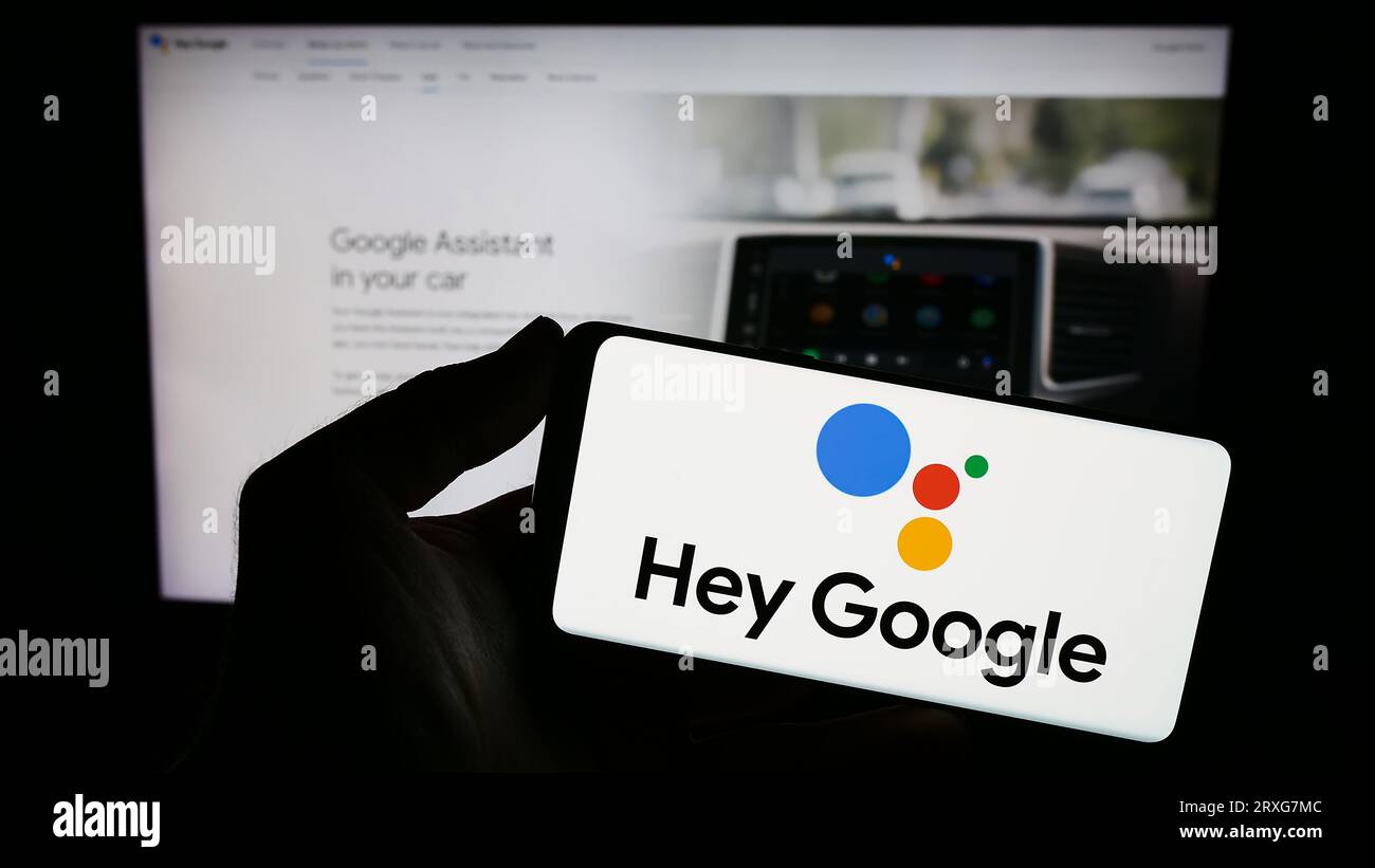 Persona che possiede uno smartphone con il logo del software di assistenza virtuale Google Assistant sullo schermo davanti al sito Web. Concentrarsi sul display del telefono. Foto Stock