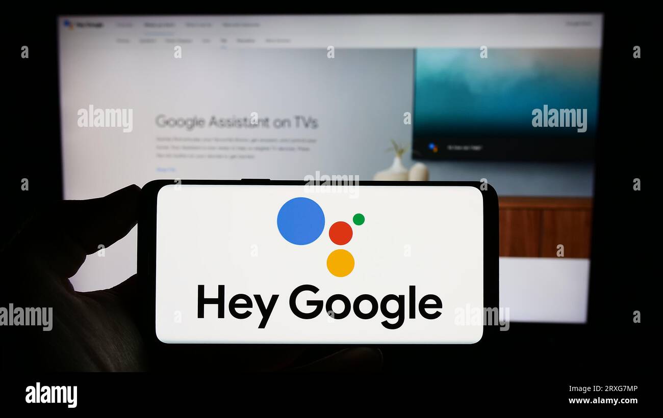 Persona che possiede un telefono cellulare con il logo del software di assistenza virtuale Google Assistant sullo schermo davanti alla pagina Web. Concentrarsi sul display del telefono. Foto Stock