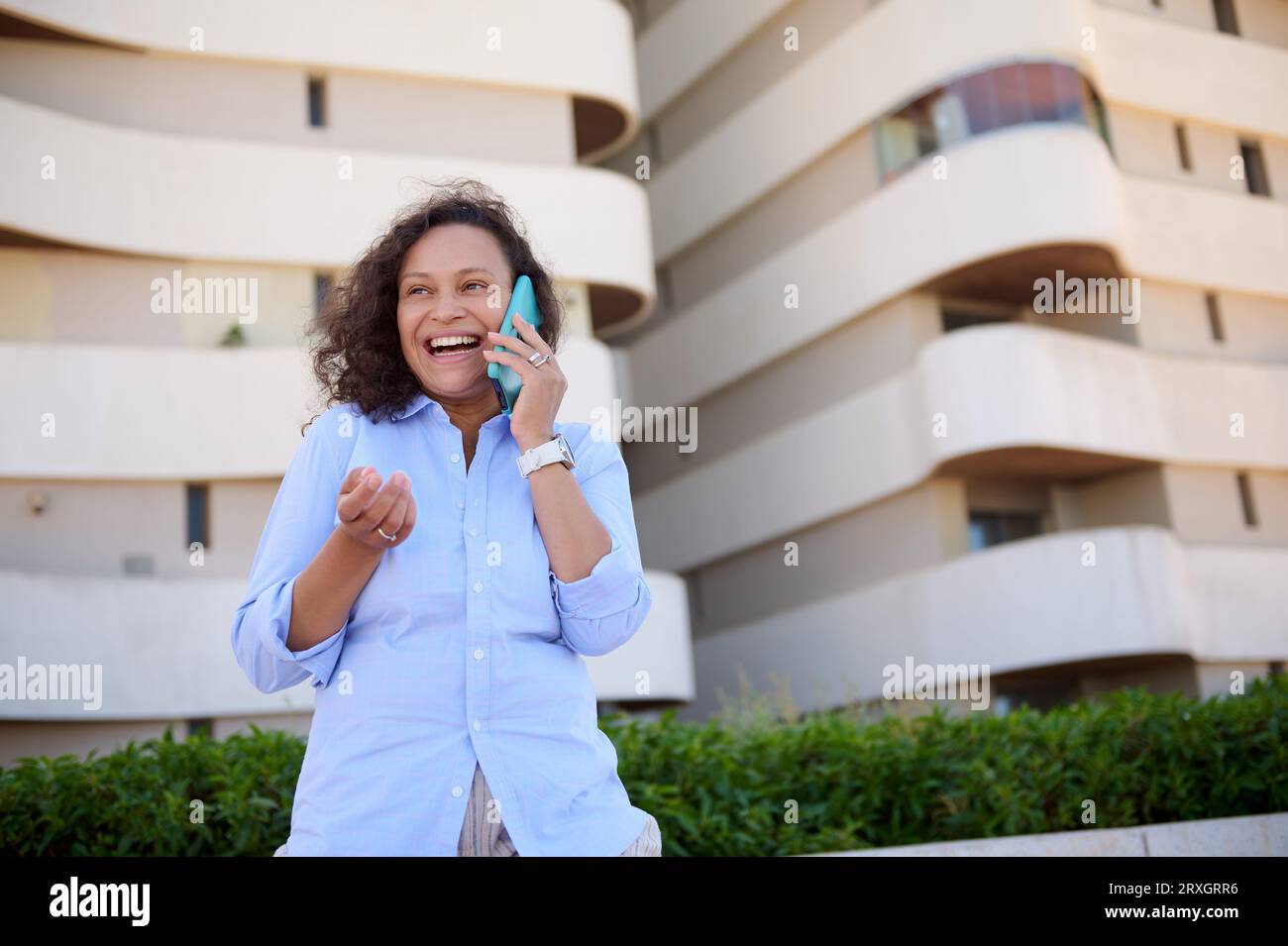Autentica donna mediorientale sicura di sé, imprenditrice di 35-39 anni in camicia casual blu, sorridente mentre parla al cellulare, contro il bui moderno Foto Stock