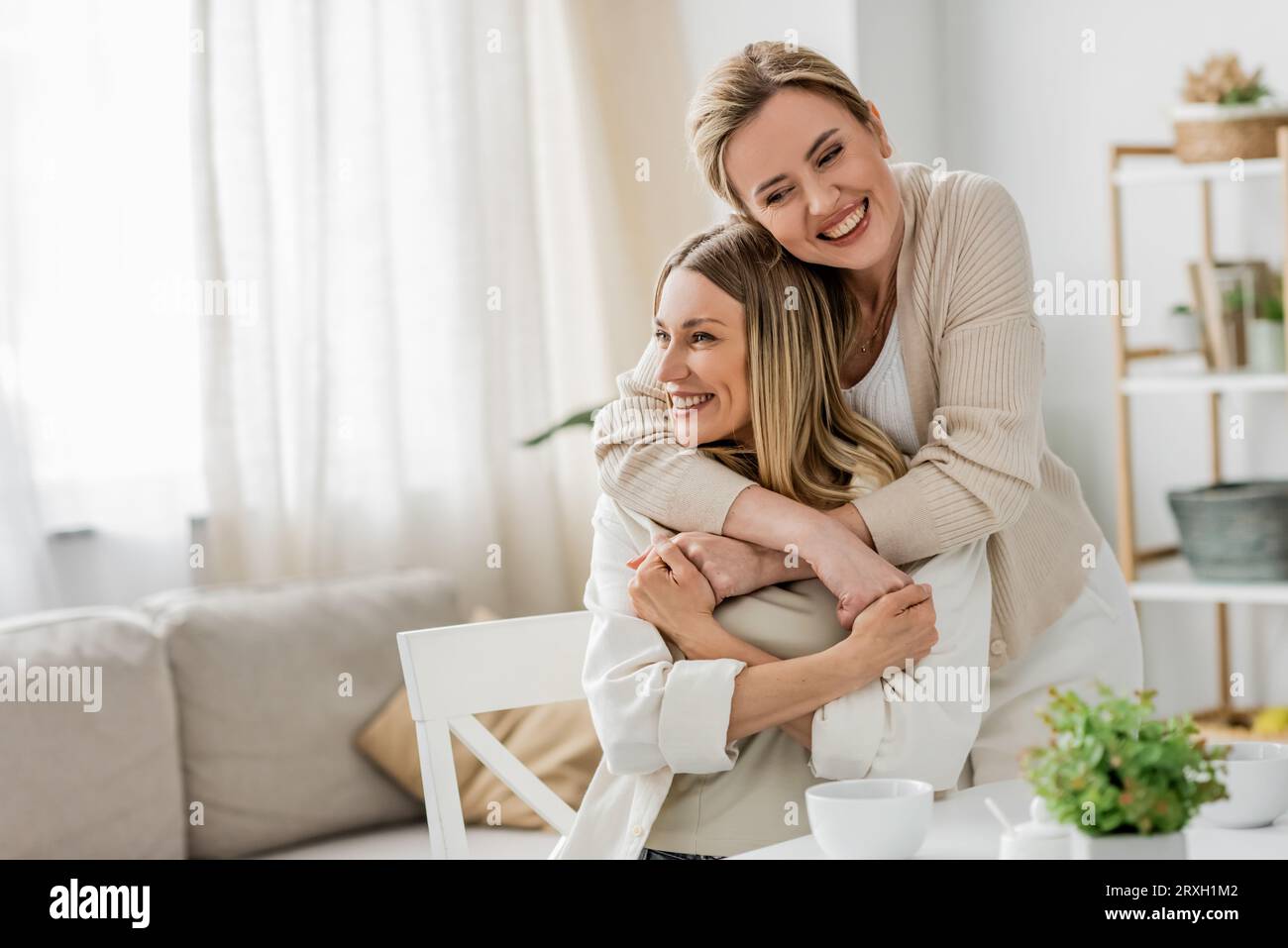 bellissime sorelle in un abbigliamento alla moda che abbracciano e sorridono sinceramente sullo sfondo della cucina, legando Foto Stock