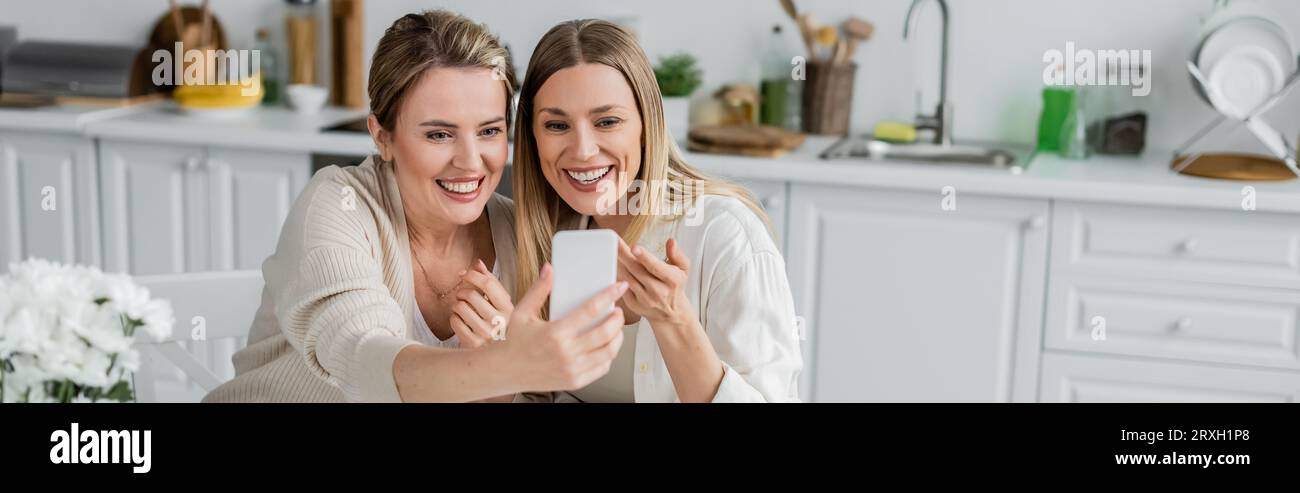 due sorelle bionde in un bell'abbigliamento che fanno selfie sullo sfondo della cucina, legano la famiglia, striscione Foto Stock