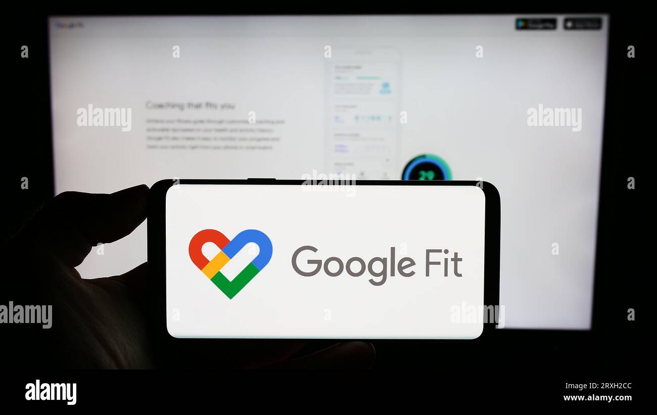 Persona che tiene il cellulare con il logo del software di monitoraggio della salute Google Fit sullo schermo di fronte alla pagina Web aziendale. Concentrarsi sul display del telefono. Foto Stock