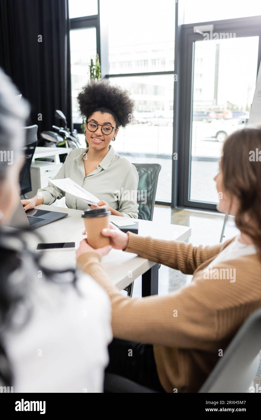 felice donna d'affari afroamericana che parla con una collega donna con un caffè per andare in un ufficio moderno Foto Stock