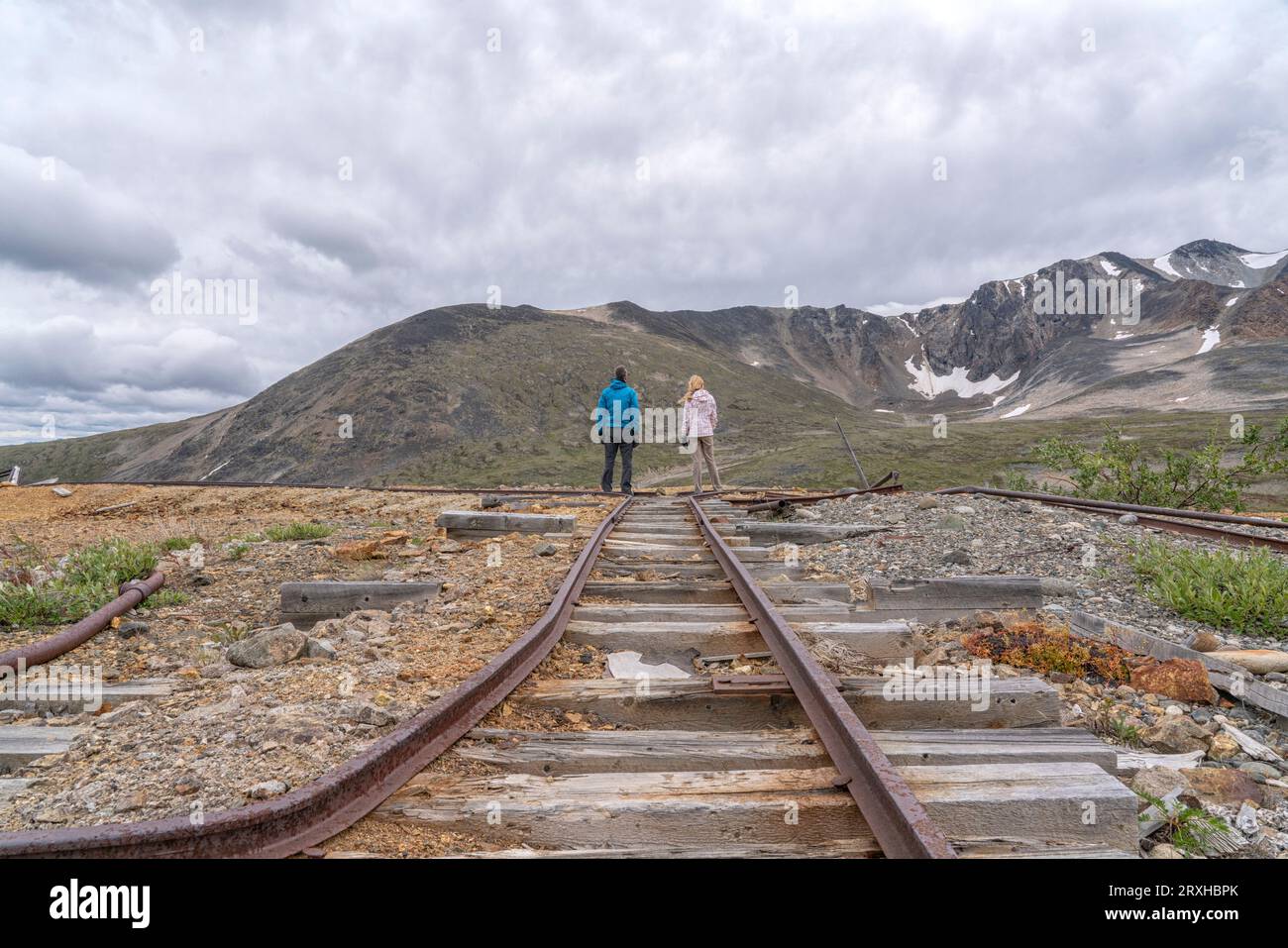 Vista posteriore di due persone in piedi su una vecchia linea ferroviaria mineraria mentre si divide con le colline del Montana Mountain intorno a loro vicino a Carcross, Yuko... Foto Stock