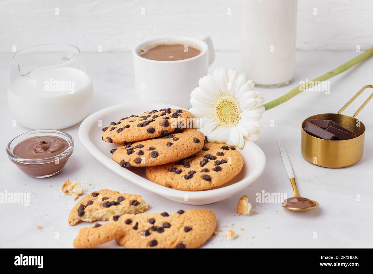 Piatto di gustosi biscotti con gocce di cioccolato, latte, salsa, tazza di caffè e fiore di camomilla su sfondo bianco Foto Stock