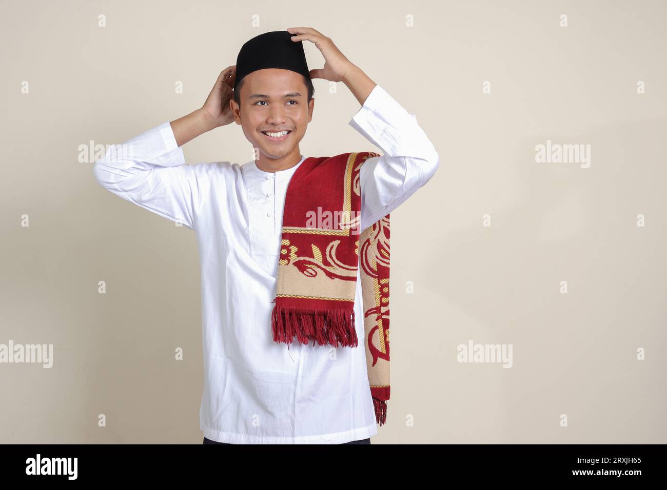 Ritratto di un attraente uomo musulmano asiatico in camicia bianca con berretto che cerca di regolare il suo songkok o skullcap nero. Immagine isolata su sfondo grigio Foto Stock