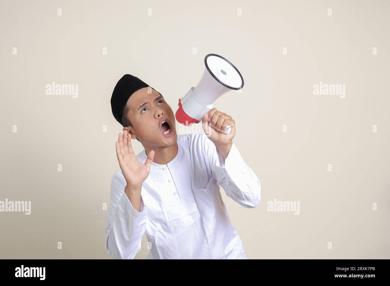 Ritratto di un attraente uomo musulmano asiatico in camicia bianca con berretto che parla più forte usando il megafono, promuovendo il prodotto. Concetto pubblicitario. Isolato Foto Stock