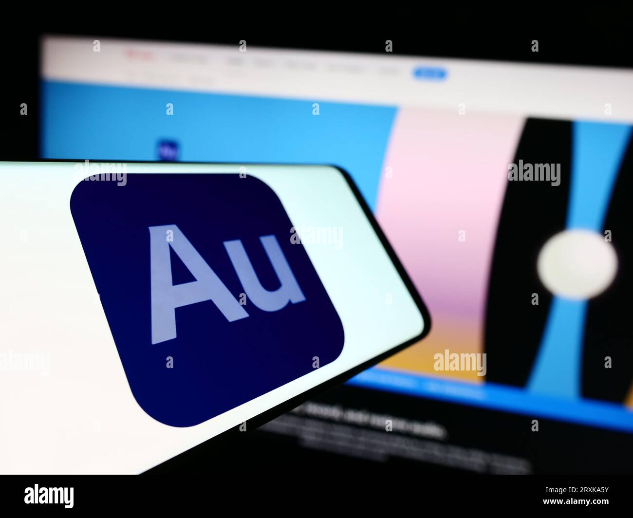 Telefono cellulare con logo della workstation audio digitale Adobe Audition sullo schermo di fronte al sito Web aziendale. Mettere a fuoco il display centrale sinistro del telefono. Foto Stock