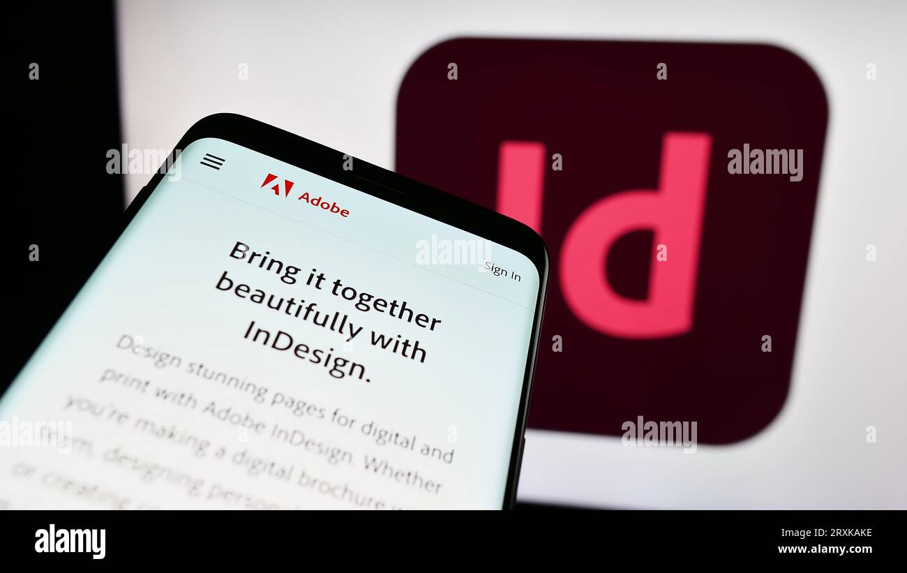Telefono cellulare con pagina Web del software di desktop publishing Adobe InDesign sullo schermo davanti al logo aziendale. Mettere a fuoco in alto a sinistra sul display del telefono. Foto Stock