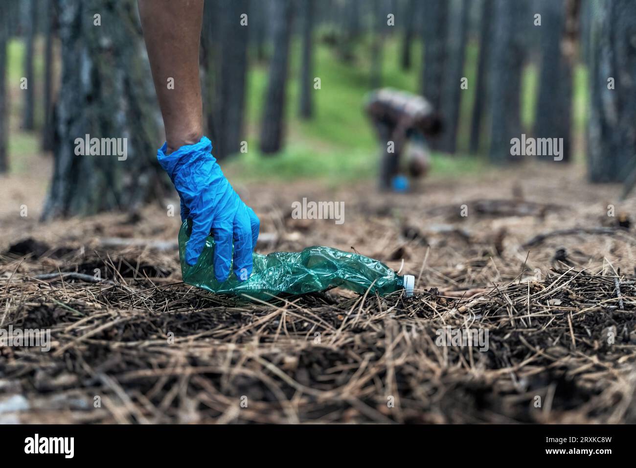 Il primo piano cattura una mano con i guanti che raccoglie un flacone di plastica scartato. Sullo sfondo, sotto il baldacchino degli alberi, un altro individuo si piega verso ga Foto Stock