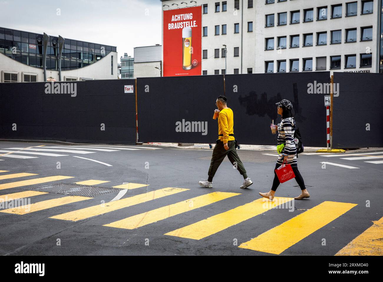 Una coppia attraversa un passaggio di fronte a una recinzione di un cantiere vicino alla cattedrale, colori e modelli di abbigliamento si adattano all'ambiente, Colonia, Foto Stock
