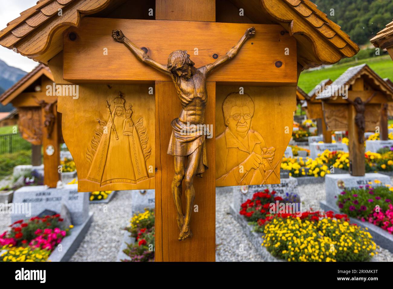 Una croce funeraria a Jaun mostra una figura radiosa di apparizione mariana nel rilievo di sinistra, forse rappresentante un sito di pellegrinaggio, e il ritratto di una donna che tiene aghi da cucito a destra, presumibilmente uno dei suoi hobby. Ognuna delle croci di legno scolpite individualmente nel cimitero dietro la chiesa di San Stefano porta un tale rilievo che raffigura la professione o l’hobby della persona sepolta sotto di essa, una tradizione iniziata nel 1948 dallo scultore autodidatta Walter Cottier e continuata dal 1988 da suo cugino Reynold Boschung. Aiuole di fiori rossi e gialli incorniciano la croce, cantone di Friburgo, Schwitze Foto Stock