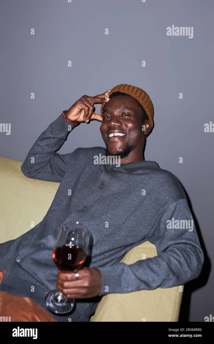 Ritratto verticale di un giovane uomo nero sorridente con vino rosso seduto sul divano sorridendo alla macchina fotografica nel nightclub Foto Stock