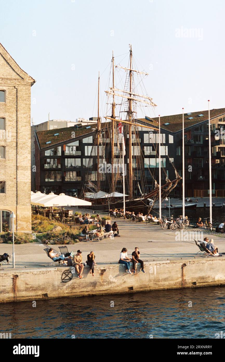 Il vecchio mondo incontra il mondo moderno a Copenaghen: Vista sulla strada, vecchia nave ed edifici moderni, persone sedute in riva al mare, che si godono il tramonto Foto Stock