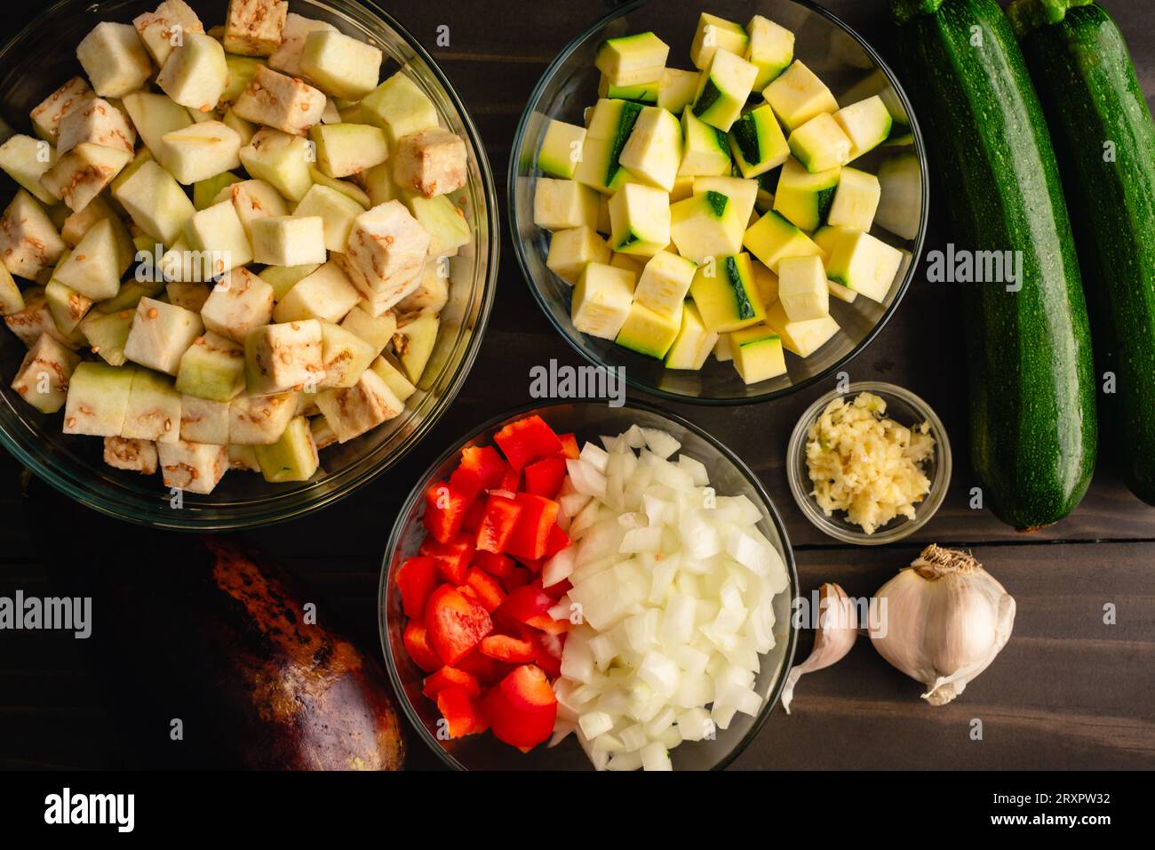 Verdure estive tritate in ciotole di vetro: Melanzane preparate, zucchine, peperone rosso, cipolla e aglio su un tavolo di legno Foto Stock