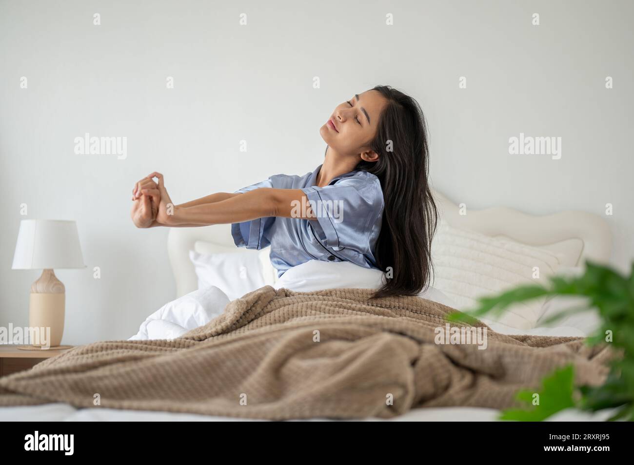 Una donna asiatica rilassata e spensierata in pigiama si sta allungando le braccia dopo essersi svegliata la mattina sul suo comodo letto. riposante, dormi bene, felici morni Foto Stock