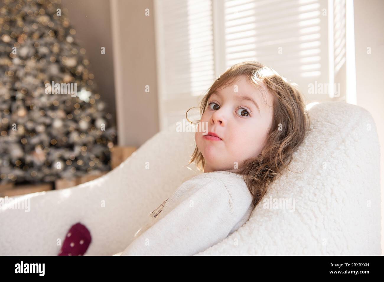 Simpatica bambina giace su una morbida sedia bianca, ingannando, facendo facce vicino all'albero di Natale. Bambino in un'atmosfera accogliente a casa. Interi festivi Foto Stock