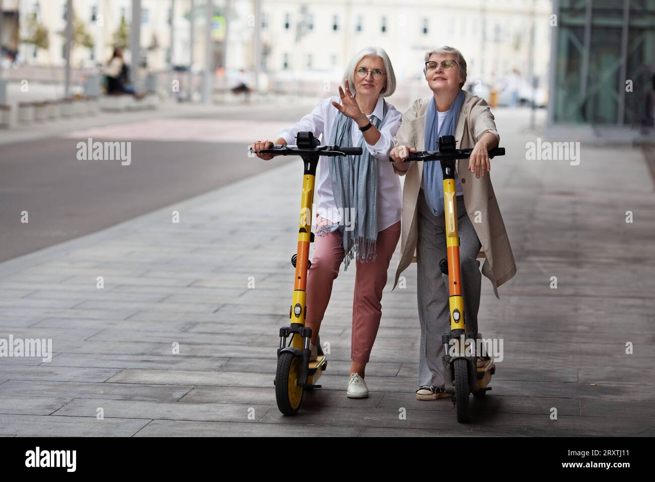 Donne anziane con i capelli grigi che si divertono a cavalcare insieme su scooter elettrici all'aperto per strada. Vecchie amiche che parlano e viaggiano per la città. Foto Stock