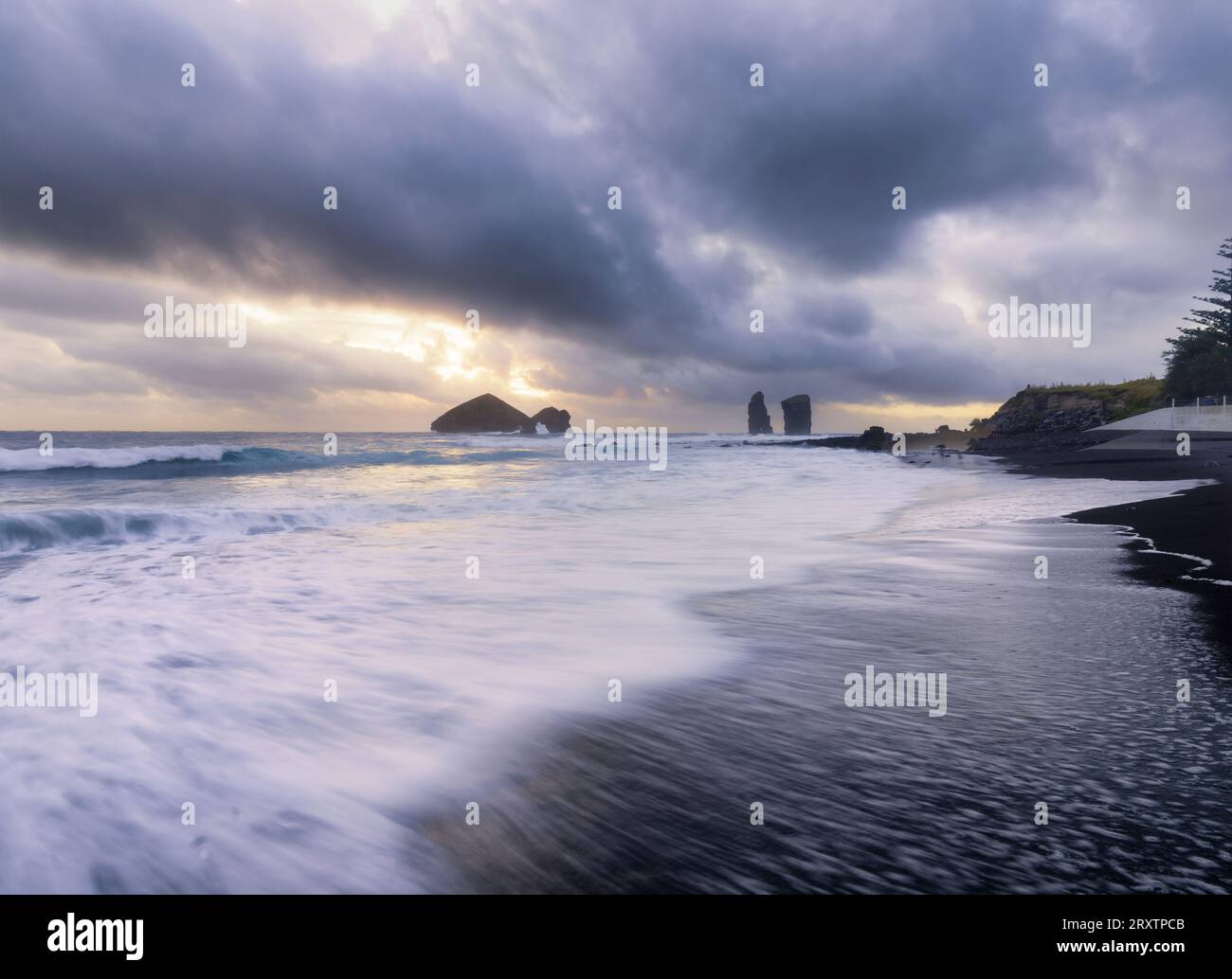 Lunga esposizione di onde sulla spiaggia di sabbia nera di Mosteiros con un tramonto nuvoloso e le pile di mare sullo sfondo, Mosteiros Foto Stock