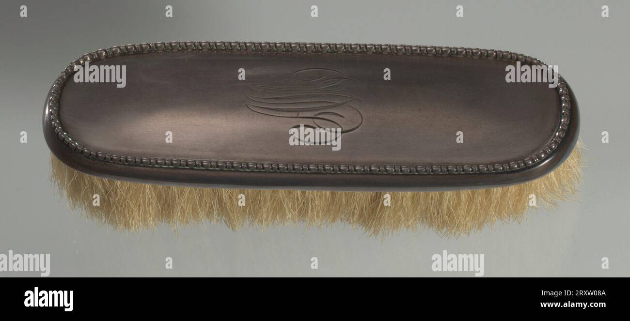 Spazzola decorativa con manico in argento. Al centro dell'impugnatura è incisa una "M" monogramma. Questa spazzola è composta da un porta pettine e da strisce di setole. Si tratta di un ovale allungato o di un rettangolo con estremità corte semicircolari. Il rivestimento esterno del manico è in argento sterling con un motivo a uovo e freccette intorno alla circonferenza del bordo superiore. L'argento copre un inserto del portabicchieri in legno che contiene setole di cavallo gialle semirigide da 1 ¿¿. Il legno è ricoperto di legno sottile o impiallacciato in plastica con fori circolari per il passaggio dei crini. L'argento è decorato in repouss¿ flo Foto Stock