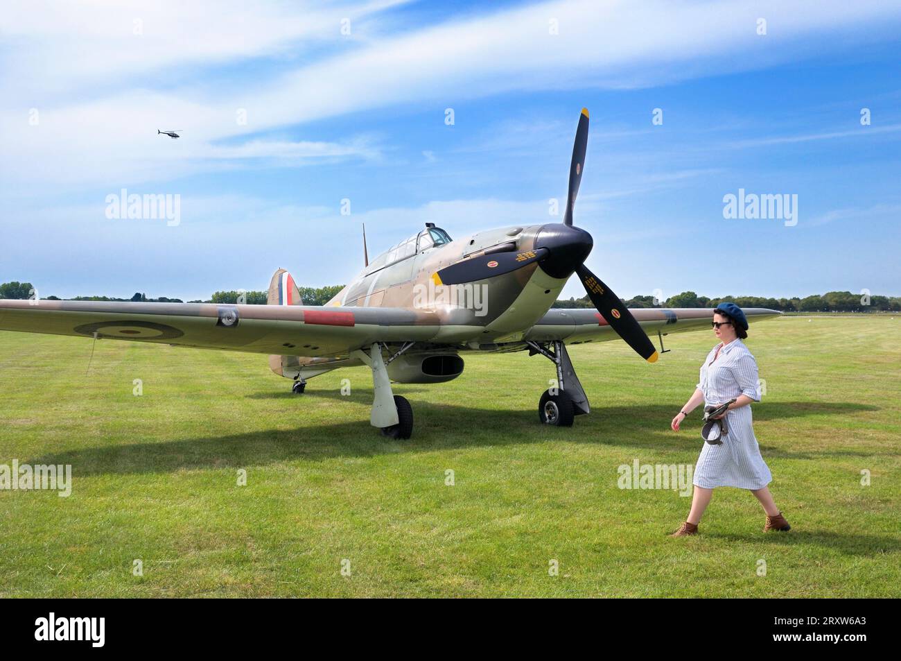 Una donna in abbigliamento d'epoca che cammina davanti a un aereo da caccia Hawker Hurricane Mk.1 P2921 GZ-L parcheggiato sull'erba al Goodwood Revival, West Sussex, Inghilterra, Regno Unito Foto Stock