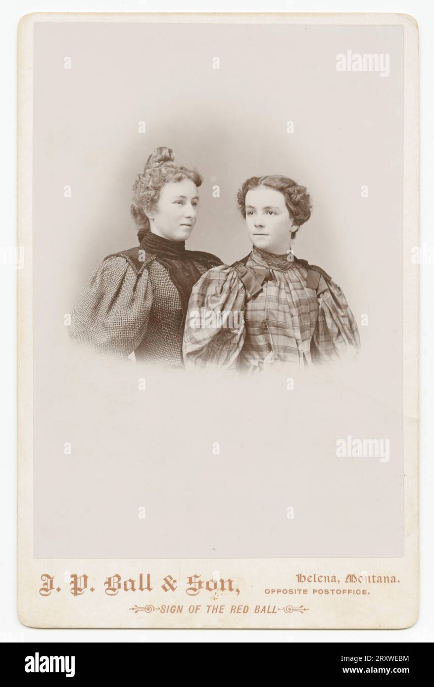 Una fotografia in bianco e nero di due donne fotografate nello studio J. P. Ball & Son di Helena, MT. La foto è montata su una scheda di colore chiaro con il nome e la posizione dello studio fotografico stampato sulla parte anteriore lungo la parte inferiore. Il ritratto è stampato in vignetta e mostra la donna dalla vita in su. La donna sul PL è posizionata proprio di fronte alla donna sul PL. Ha la testa girata e guarda oltre il telaio a destra. Indossa un abito a quadri con fiocchi alle spalle. La donna delle PR indossa un abito in lana scura con colletto nero e pelo di capelli Foto Stock