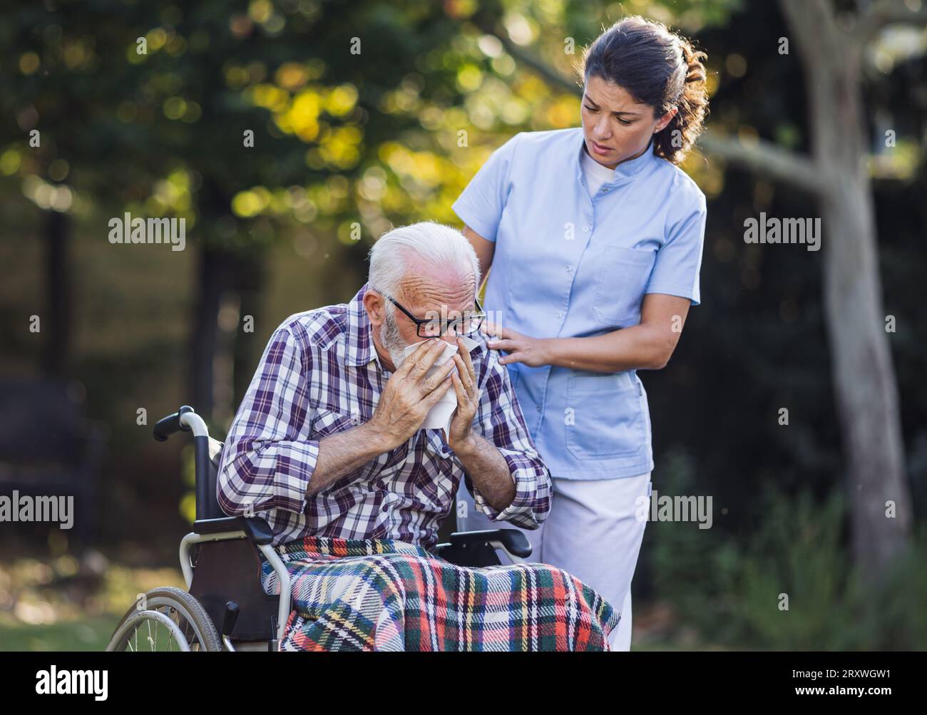 Uomo anziano in sedia a rotelle che starnutisce nel tovagliolo mentre la giovane infermiera si prende cura di lui nel parco Foto Stock