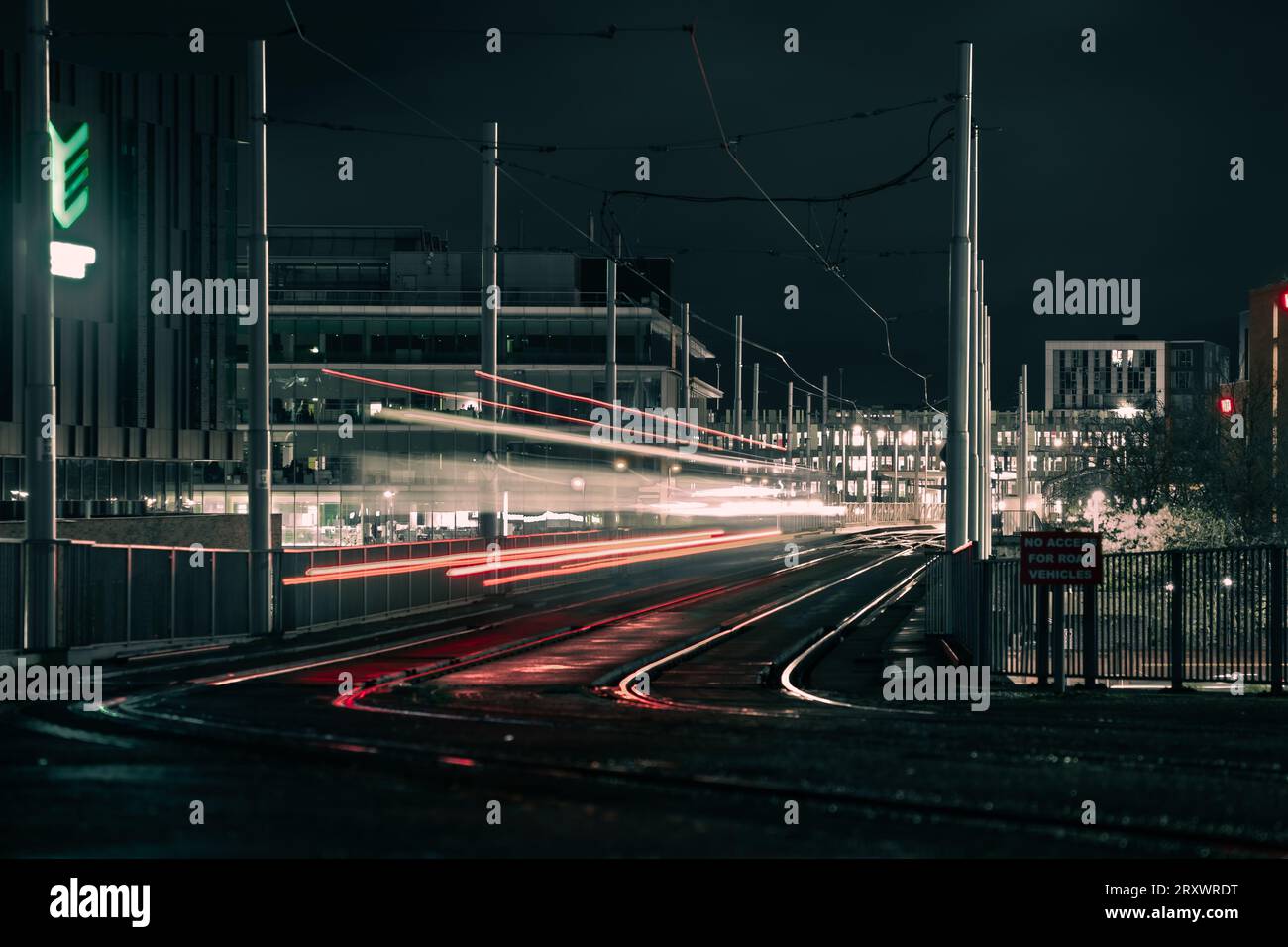 Movimento offuscato dei tram che viaggiano su rotaie a Nottingham City di notte con sentieri leggeri e edifici illuminati sullo sfondo della città Foto Stock